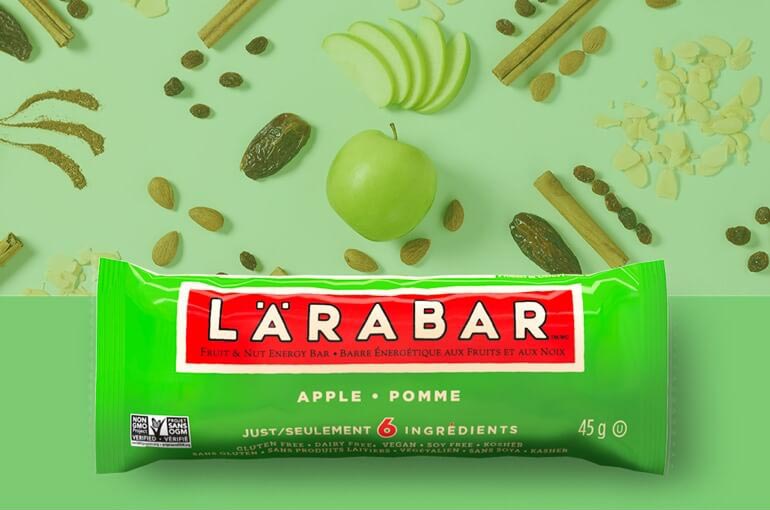 Une barre Lärabar Pomme appuyée sur des dattes, des amandes, des pommes et de la cannelle en toile de fond